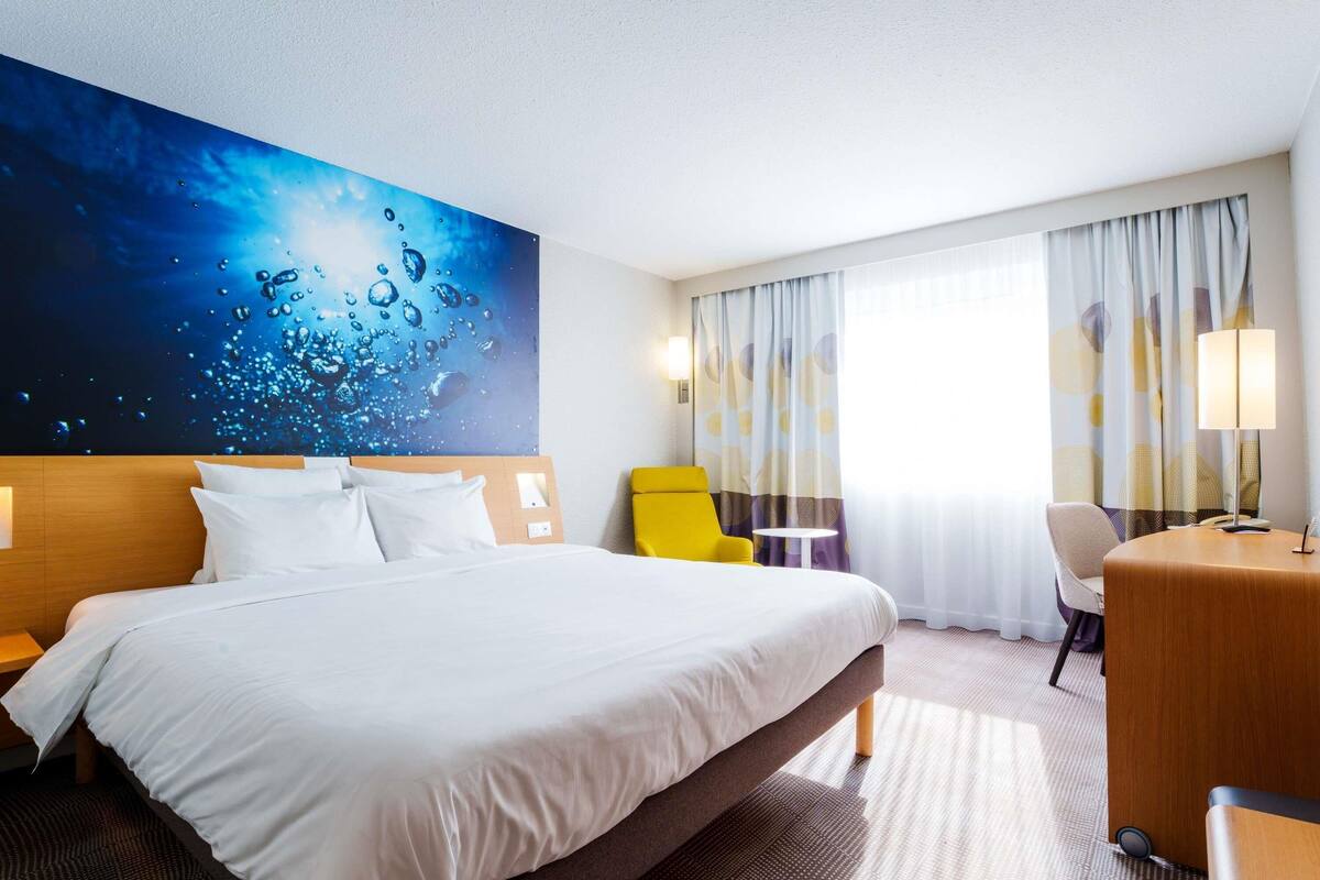 A hotel room with a large bed, a desk, a chair, a small table, and a wall with a water-themed artwork.