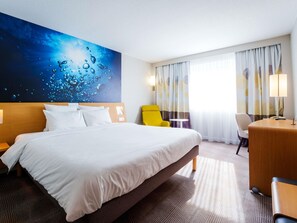 Habitación de hotel con una cama grande, un escritorio, una silla, una mesita y un cuadro de temática acuática en la pared.