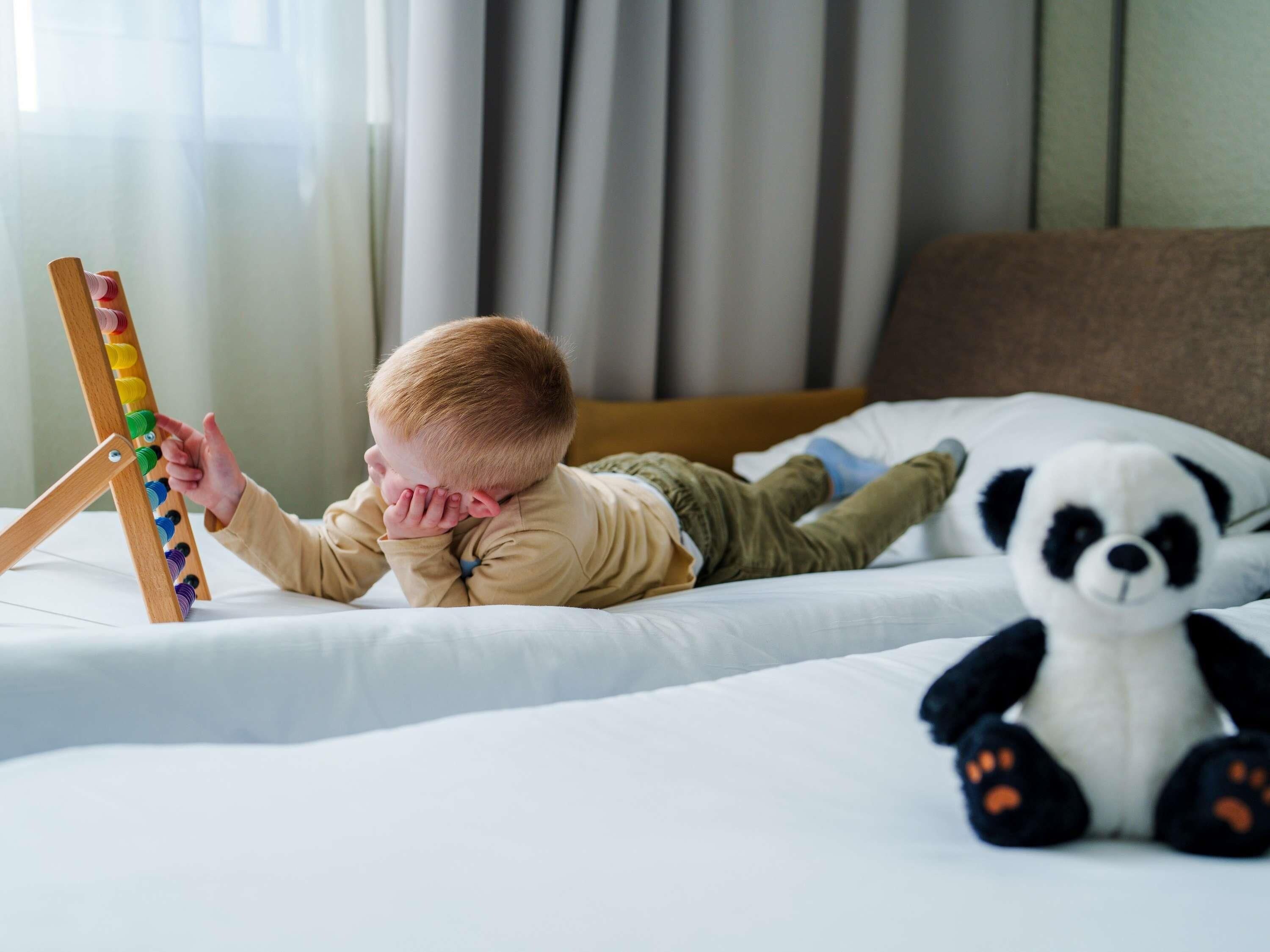 A child lying on a bed with a panda plush toy and a wooden toy structure.