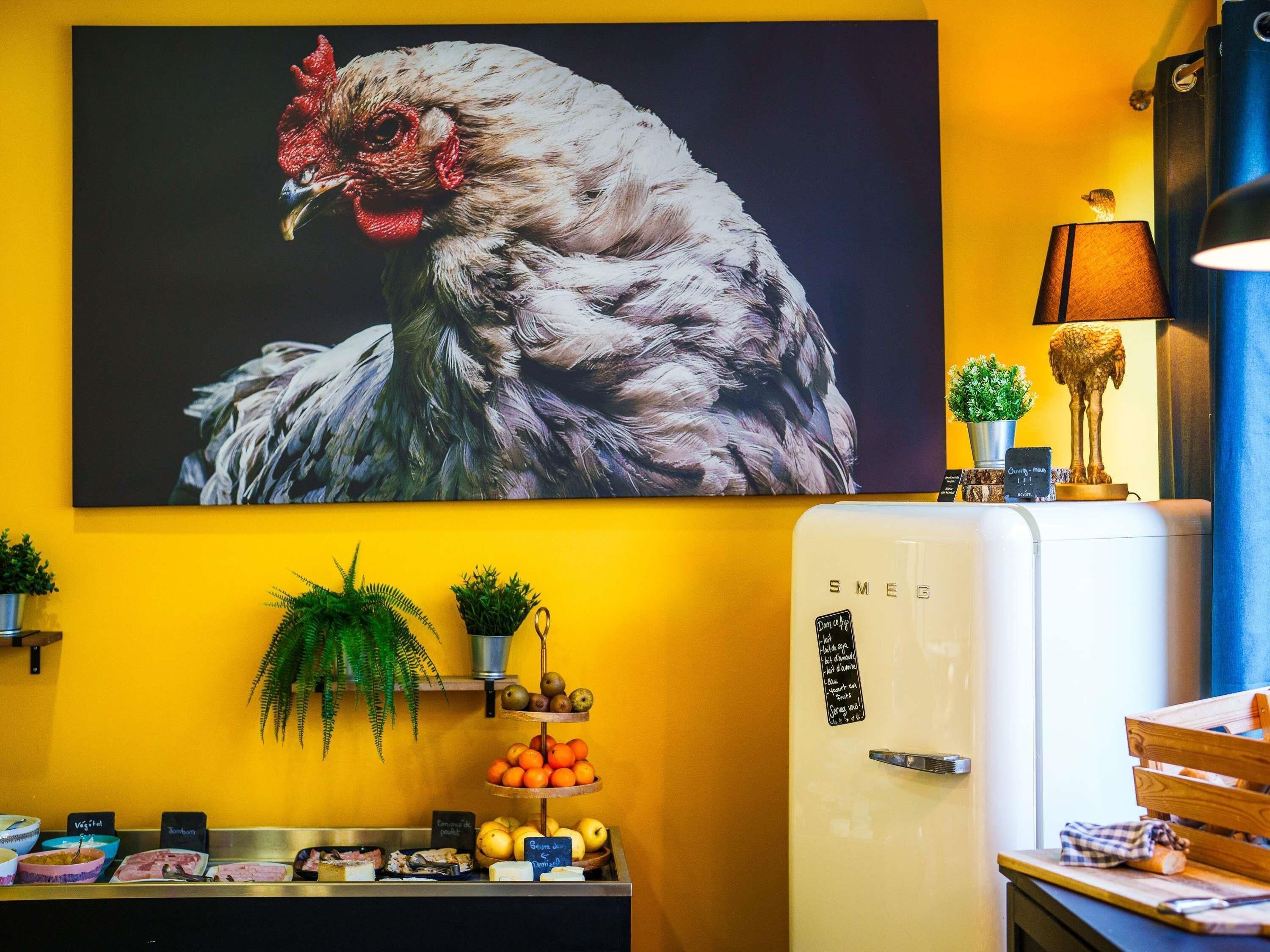 A kitchen with a refrigerator, a wall-mounted painting of a rooster, and a fruit display.
