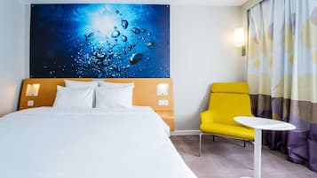 Une chambre d’hôtel avec un grand lit, un fauteuil jaune, une petite table blanche et un mur orné d’un tableau abstrait bleu.