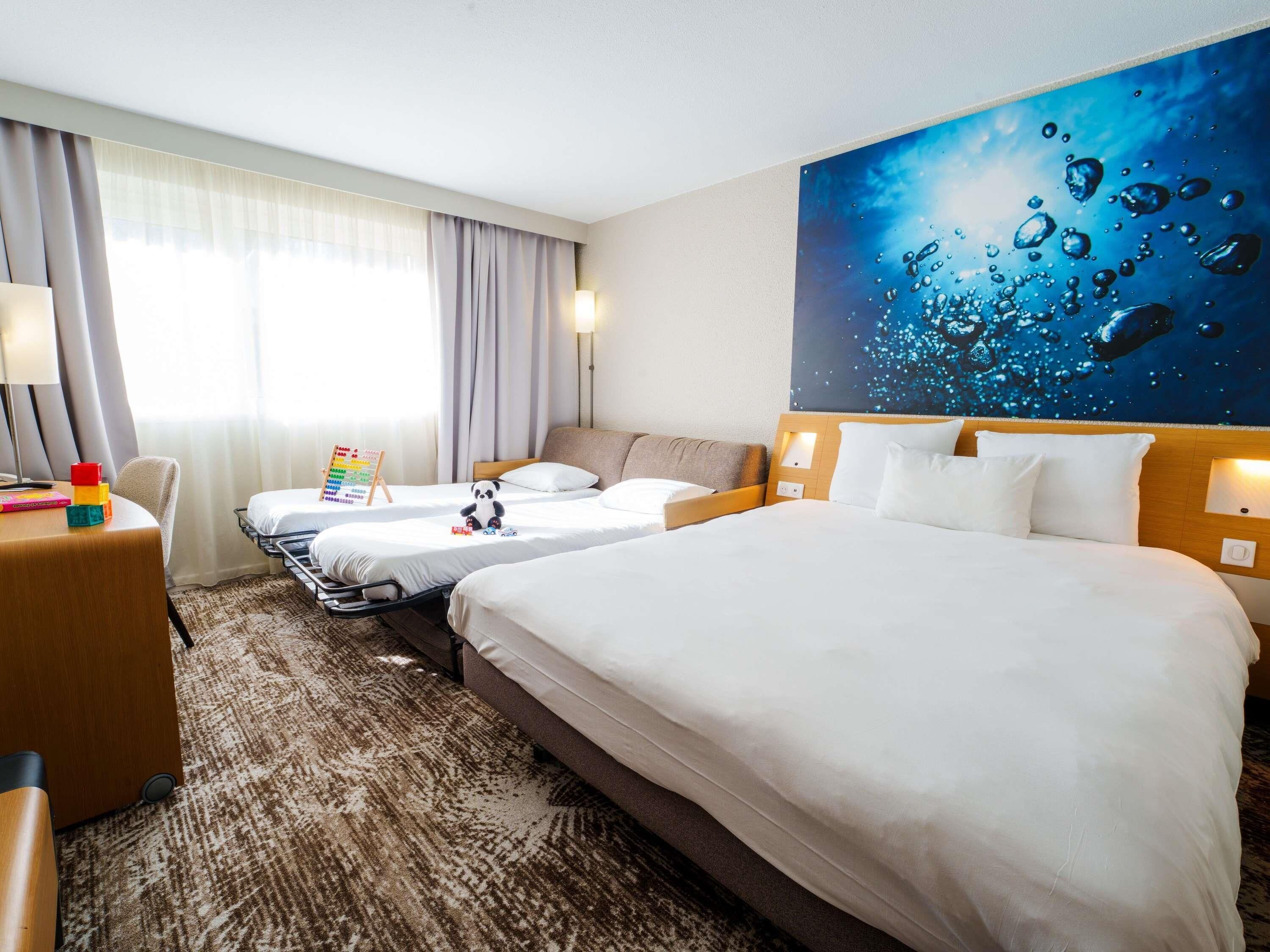 A hotel room with two beds, a desk, a lamp, and a wall painting.