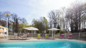 A swimming pool with lounge chairs and umbrellas, surrounded by a fence and trees. - Hôtel Novotel Saint Avold (Saint-Avold)