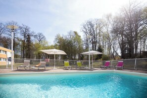A swimming pool with lounge chairs and umbrellas, surrounded by a fence and trees. - Hôtel Novotel Saint Avold (Saint-Avold)