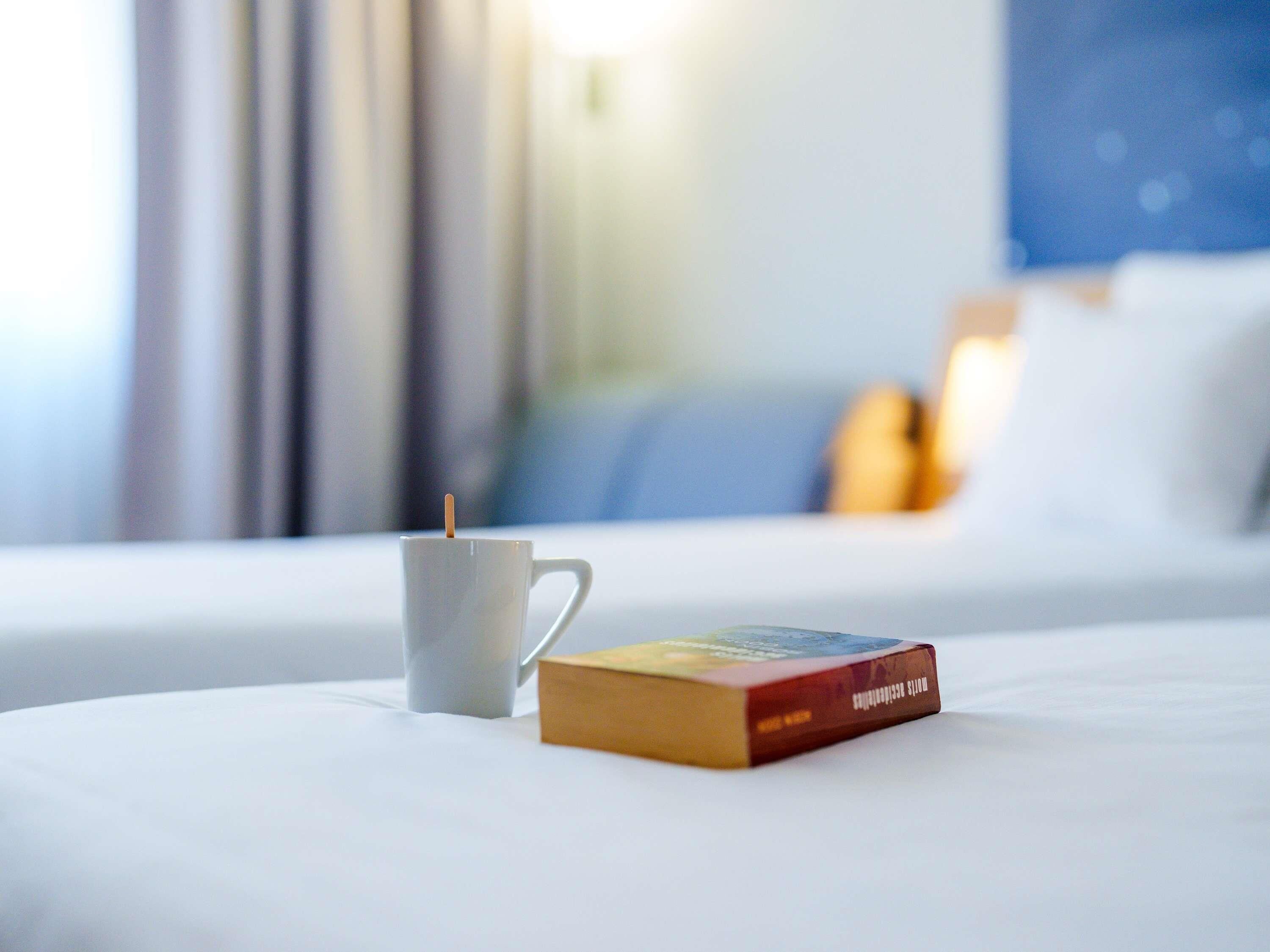 Una habitación de hotel con una cama, una taza blanca y un libro sobre la cama.