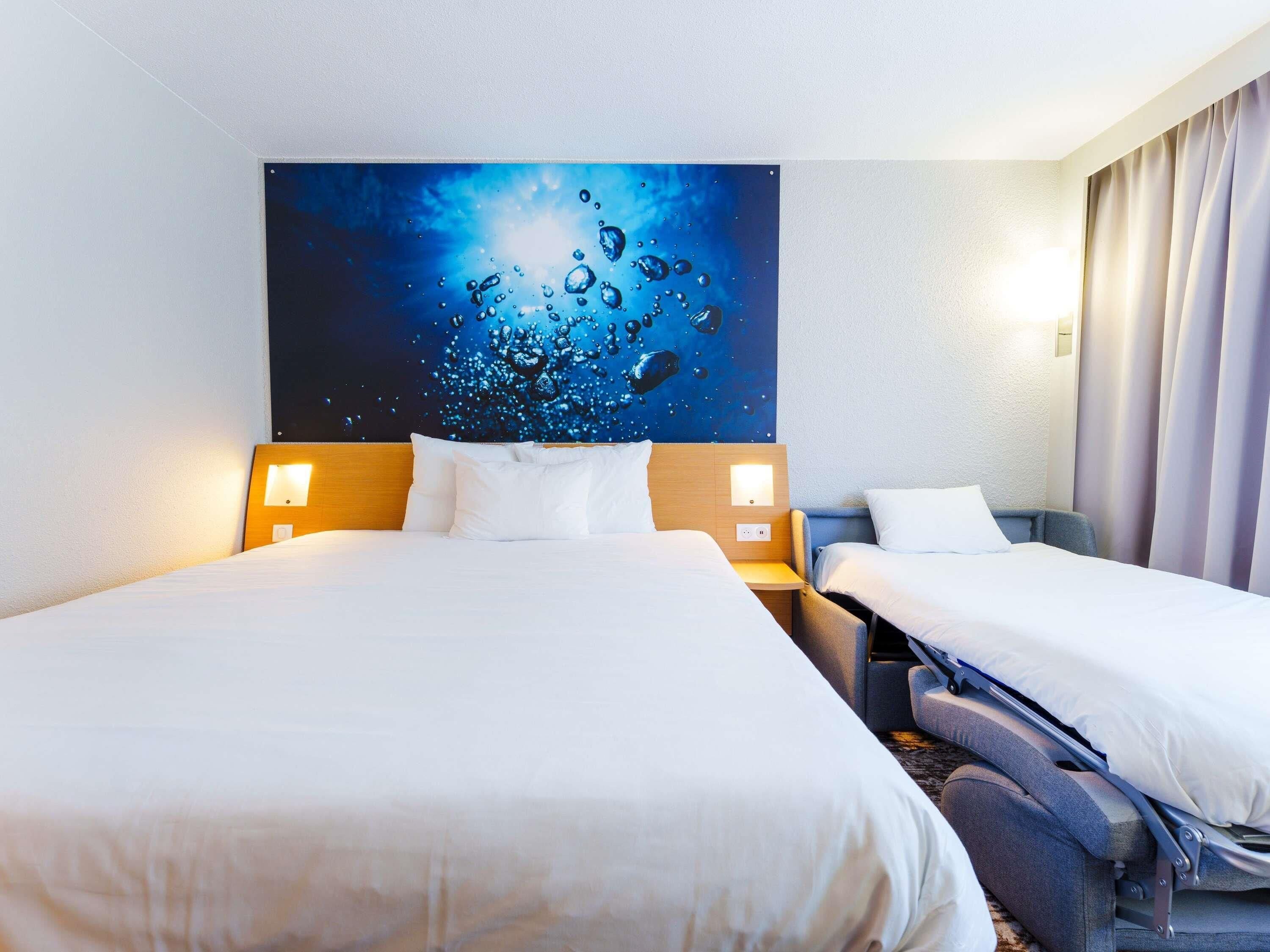 A hotel room with two beds, a large wall art of water droplets, and a nightstand with a lamp.