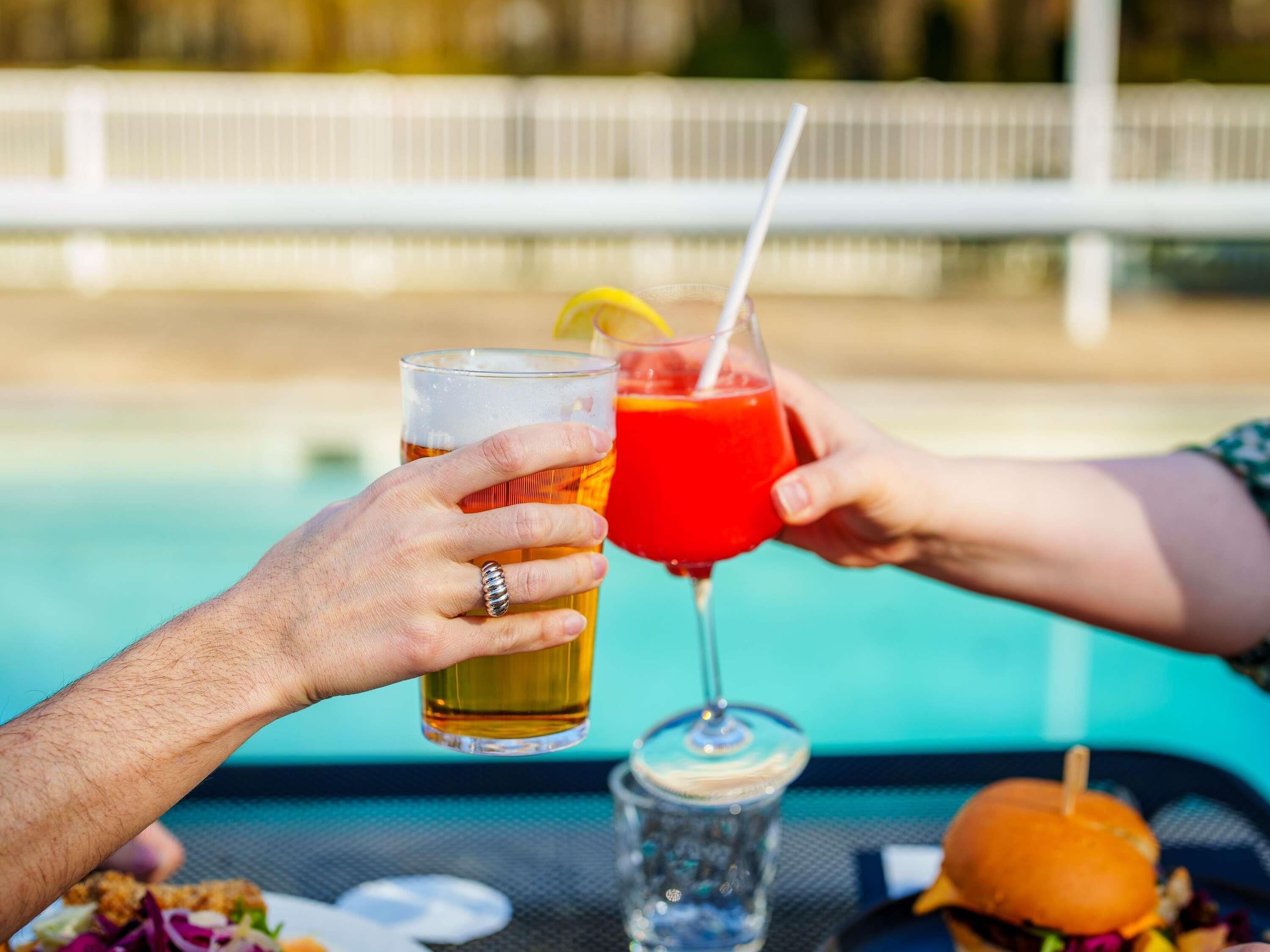 Deux personnes sont assises à une table au bord de la piscine, tenant chacune un verre, et devant elles se trouvent un hamburger et une salade.