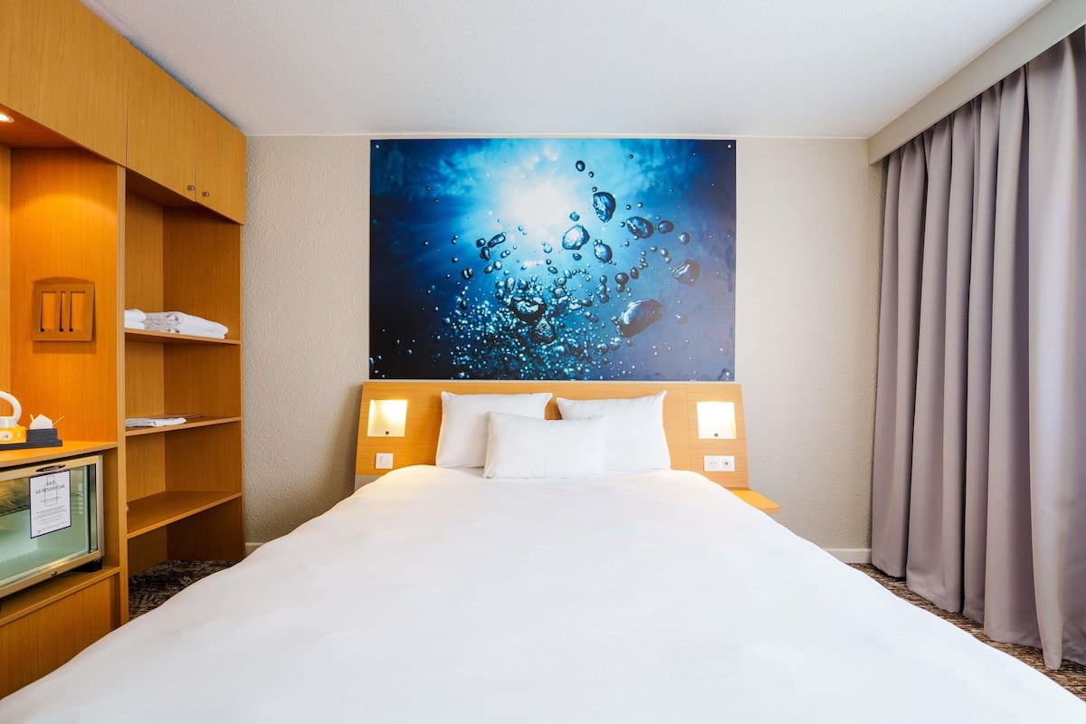 A hotel room with a large bed, a wooden wardrobe, a desk with a lamp, and a painting of water droplets on the wall.