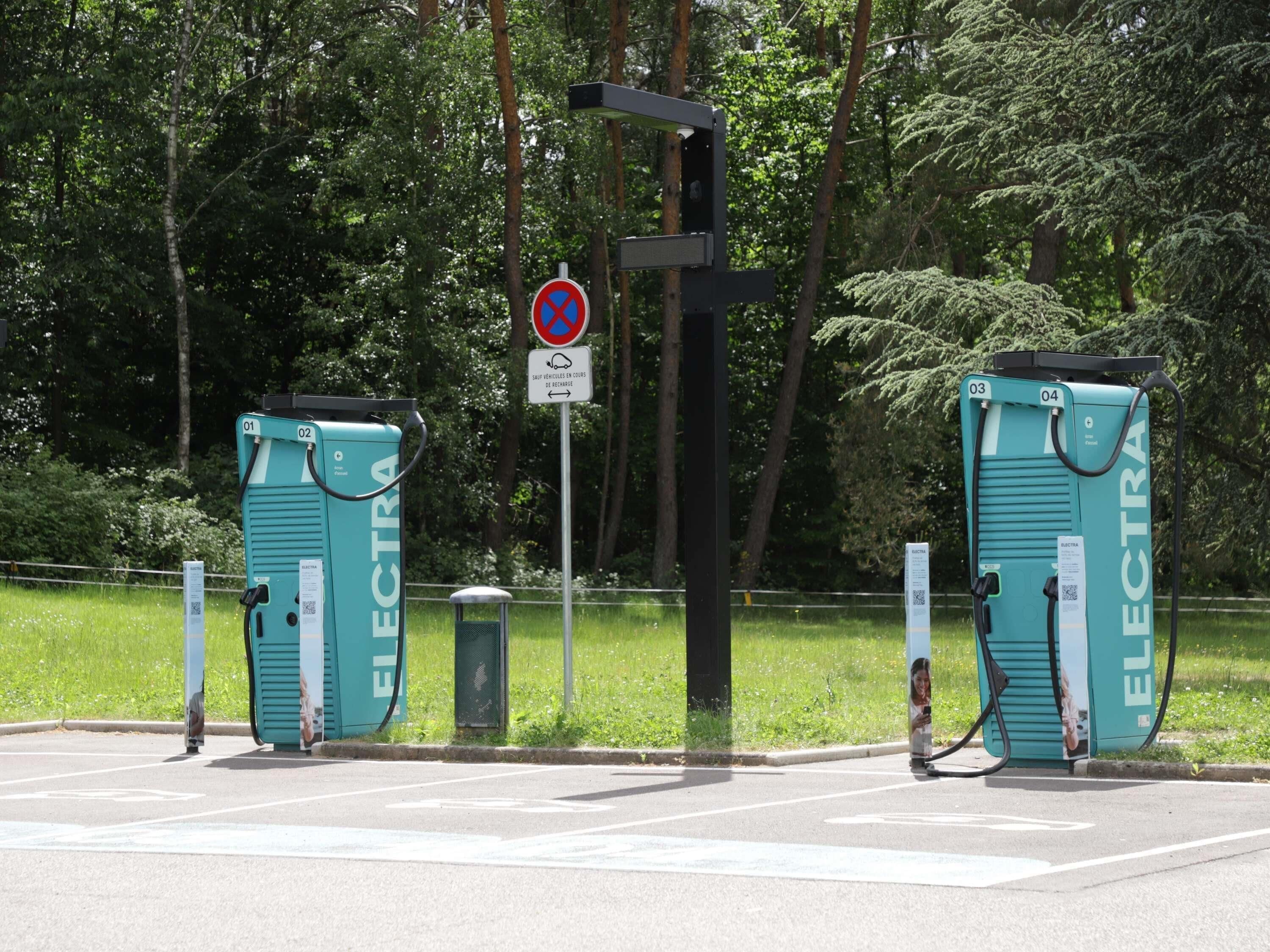 Dos estaciones de carga para vehículos eléctricos con la etiqueta 'ELECTRA', códigos QR y una señal de no estacionamiento al fondo.