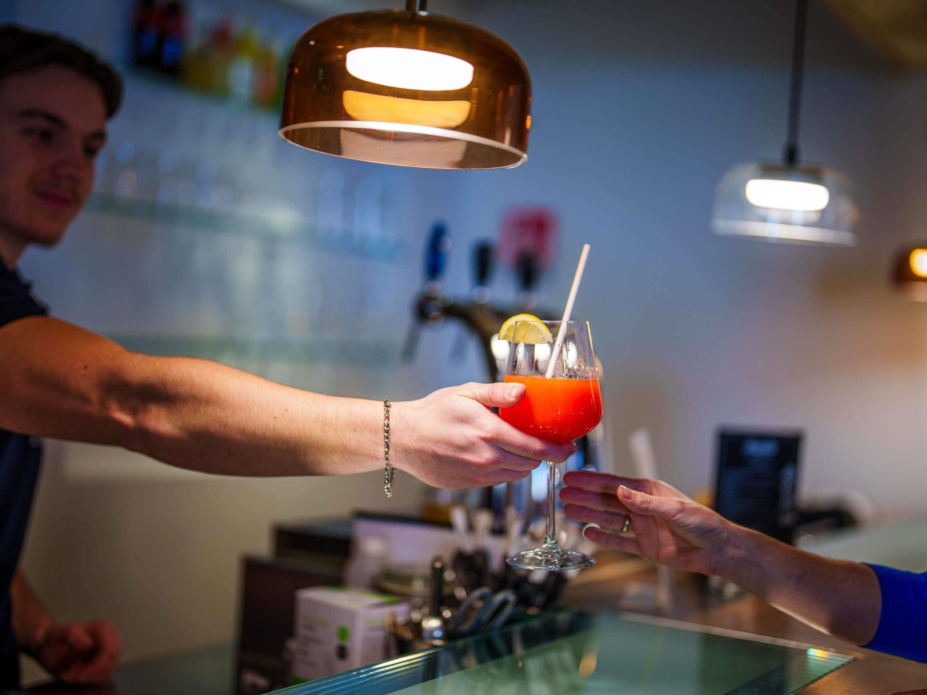 A bartender serving a cocktail to a customer at a bar with a modern design and pendant lighting.