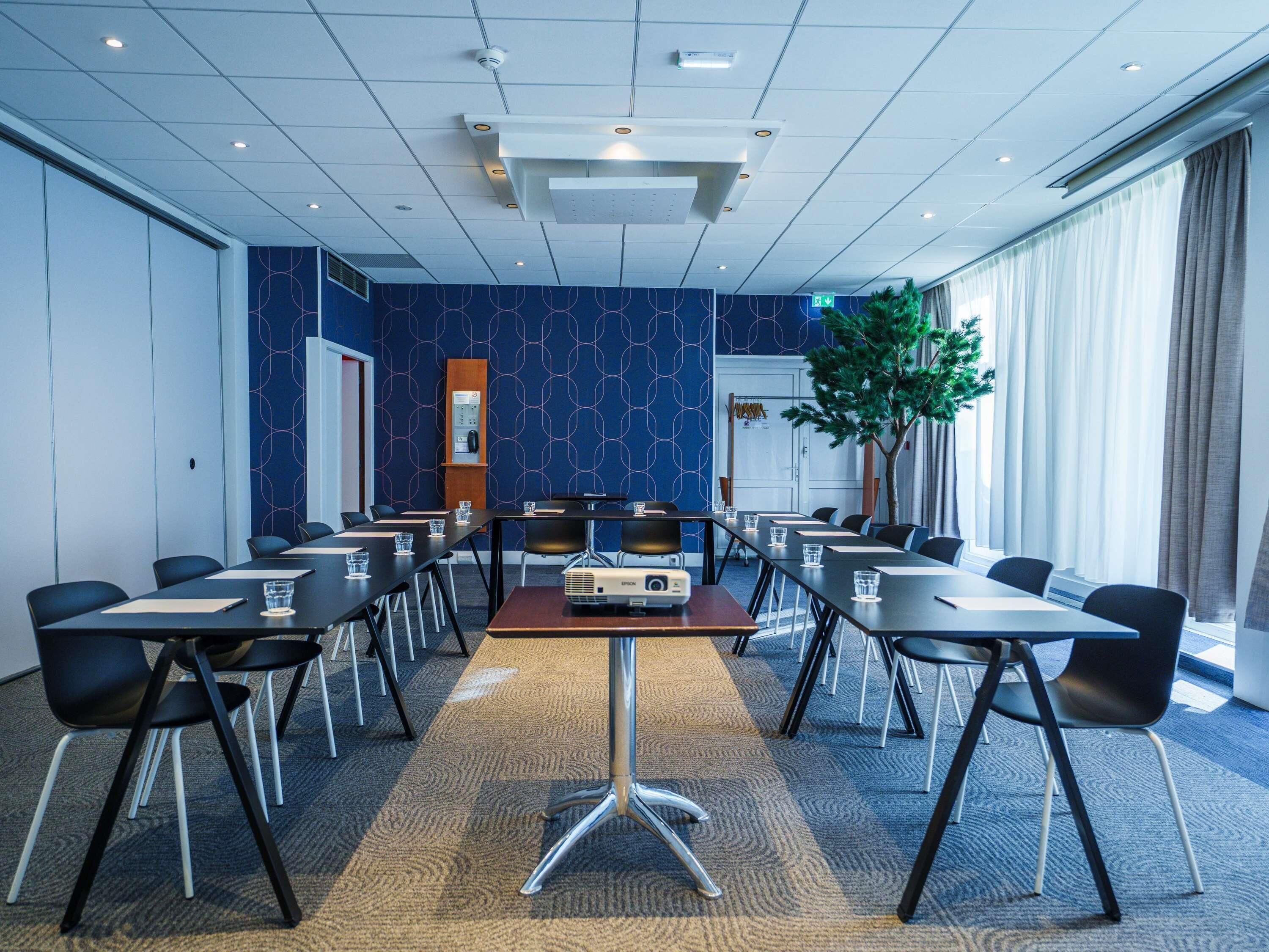 Een vergaderruimte met een lange tafel, meerdere stoelen, een projectiescherm en een groot raam met gordijnen.