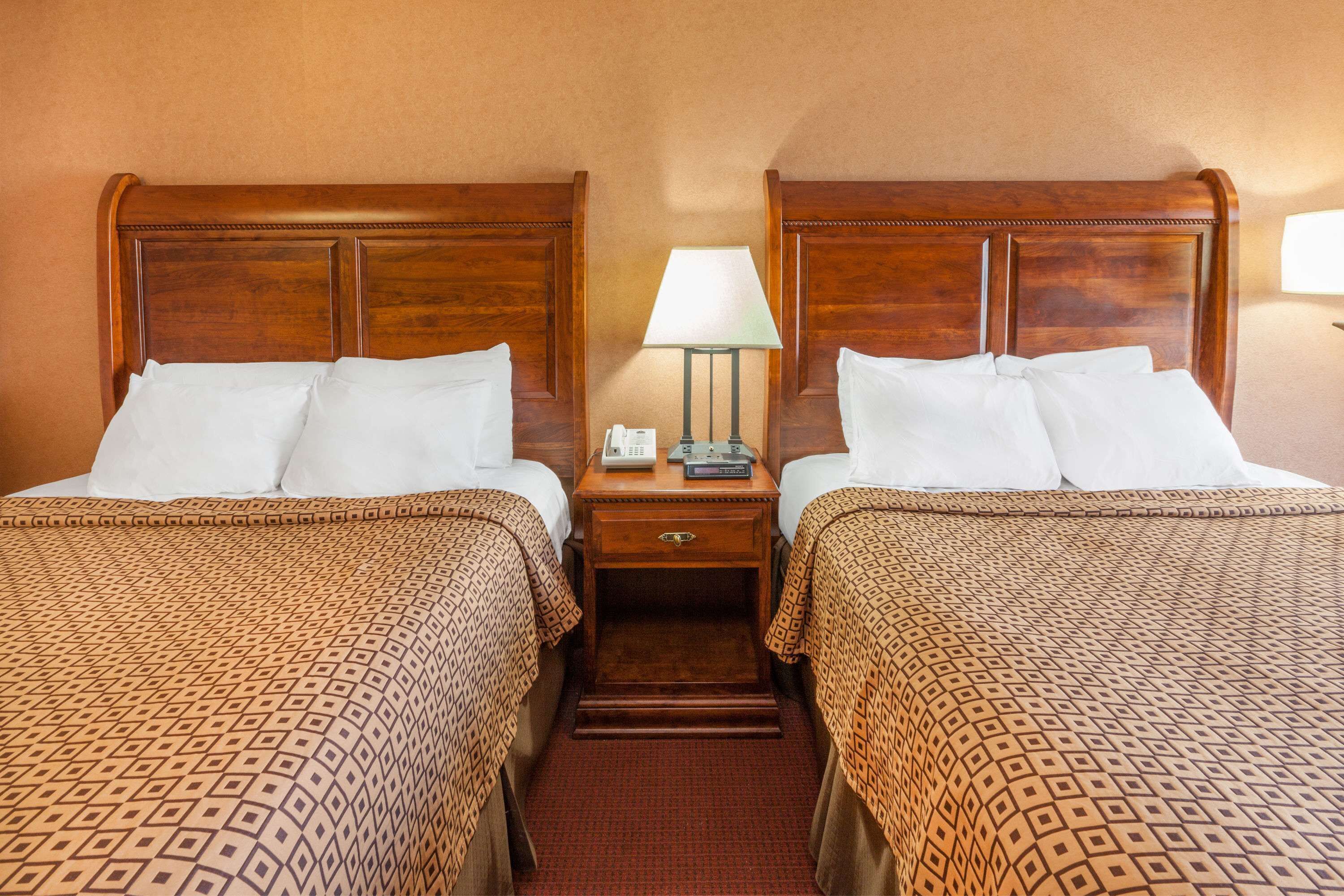 A hotel room with two beds, each with a wooden headboard, a bedside table with a lamp, and a patterned bedspread.