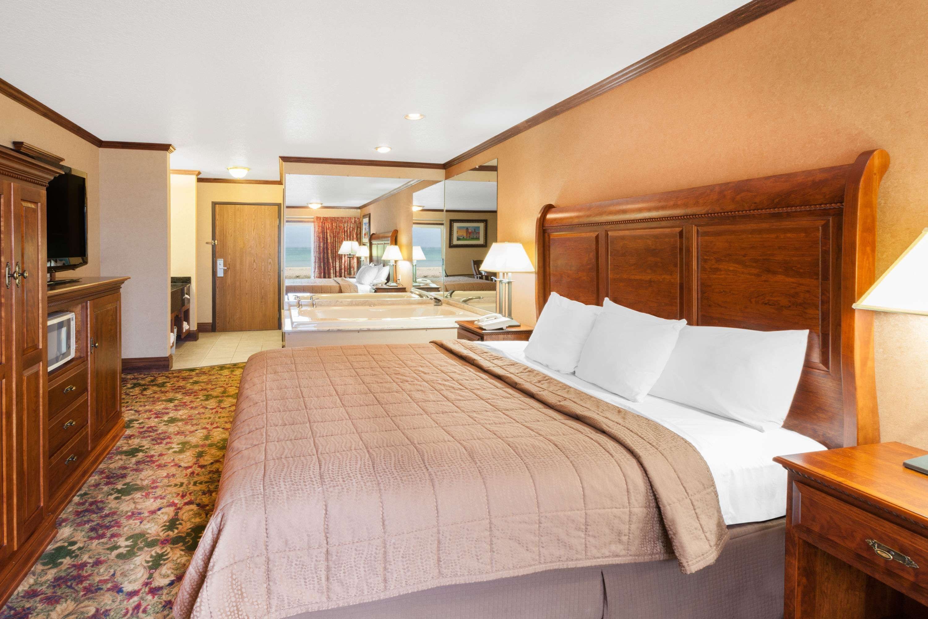 A hotel room with a large bed, wooden headboard, a television, a desk with a lamp, and a view of the beach through the door.