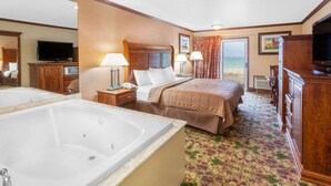 A hotel room with a large bed, a sitting area with a chair and a table, a television, a bathroom with a bathtub, and a view of the beach. - Ramada by Wyndham Mackinaw City Waterfront (Mackinaw City)