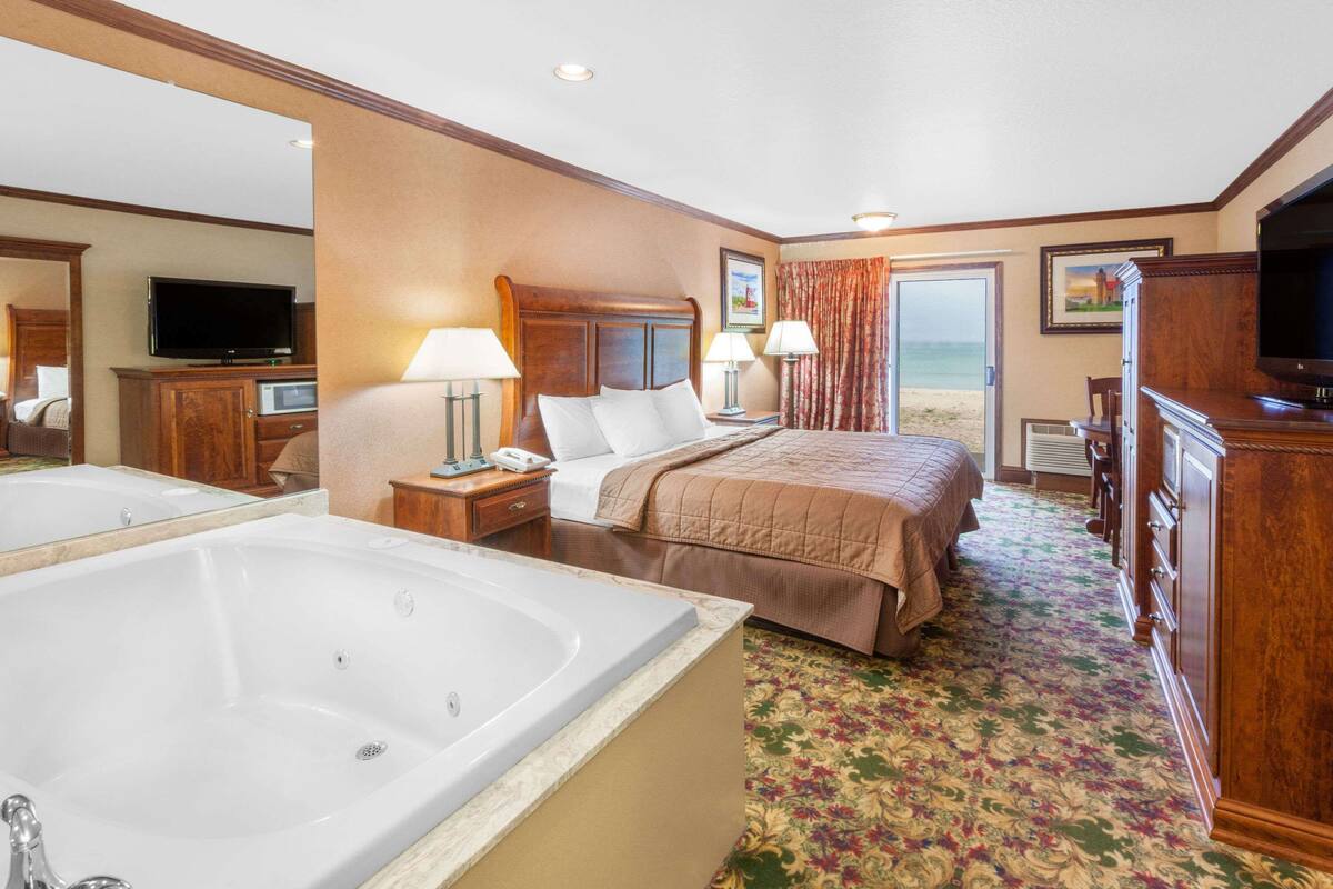 A hotel room with a large bed, a sitting area with a chair and a table, a television, a bathroom with a bathtub, and a view of the beach.
