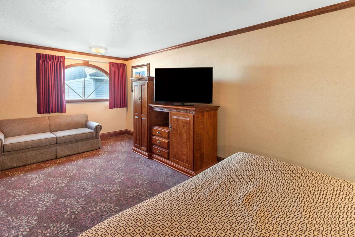 A hotel room with a sofa, a TV on a wooden cabinet, a patterned carpet, and a window with curtains.