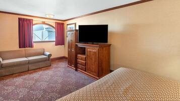 Habitación de hotel con sofá, televisor sobre un mueble de madera, alfombra estampada y ventana con cortinas.