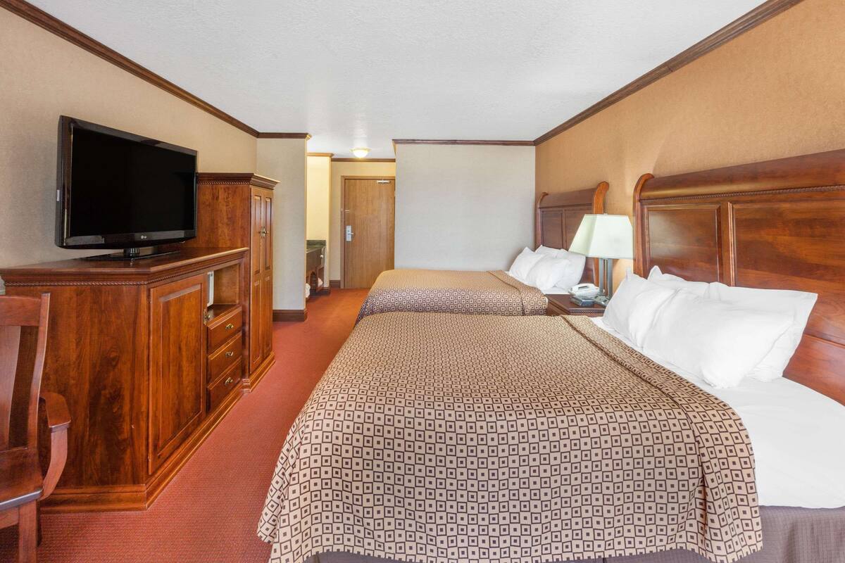 A hotel room with a large flat-screen TV, wooden furniture, a bed with patterned bedding, and a nightstand with a lamp.