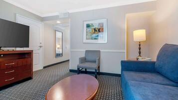 Une chambre d’hôtel équipée d’un téléviseur à écran plat, d’une table basse en bois, d’un canapé bleu, d’un fauteuil gris, d’une œuvre d’art encadrée au mur et d’une lampe sur une table de chevet.