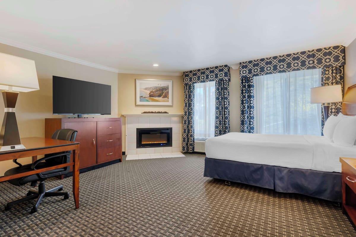 A hotel room with a bed, desk, chair, TV, fireplace, and patterned curtains.