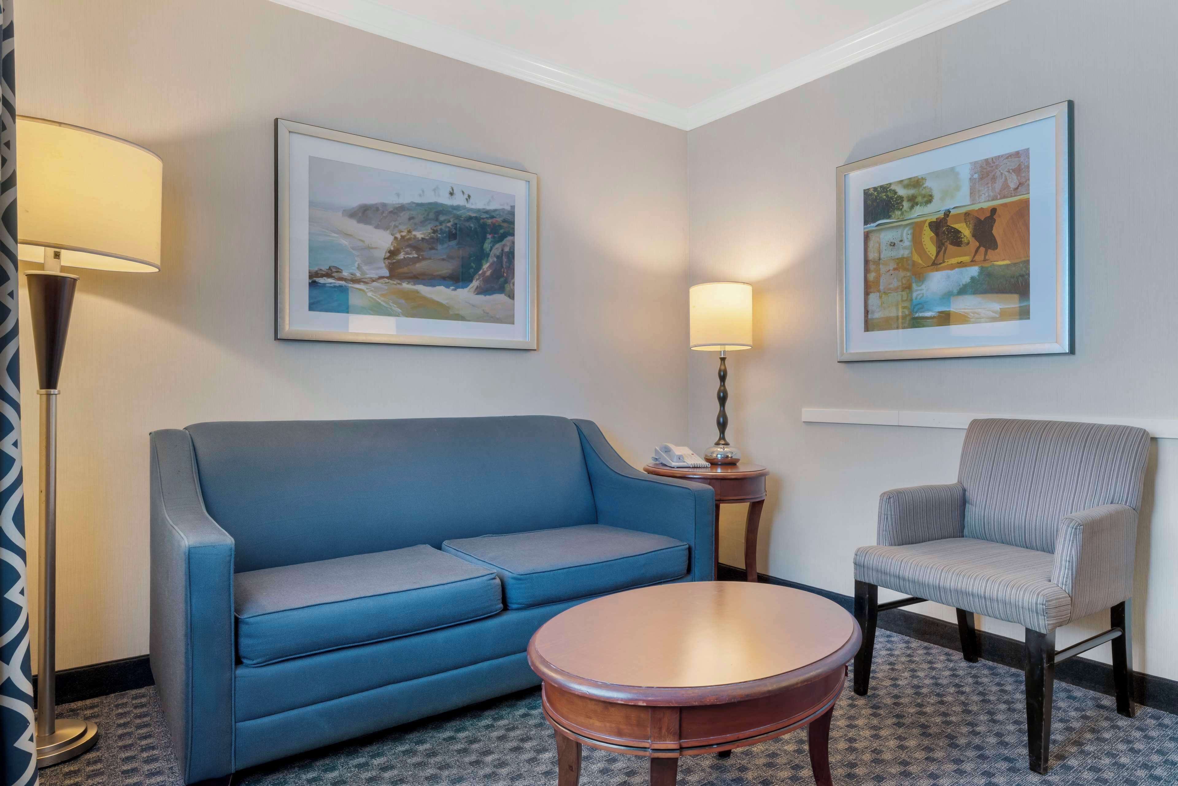 A hotel room with a blue sofa, a striped armchair, a round wooden coffee table, and two framed artworks on the walls.