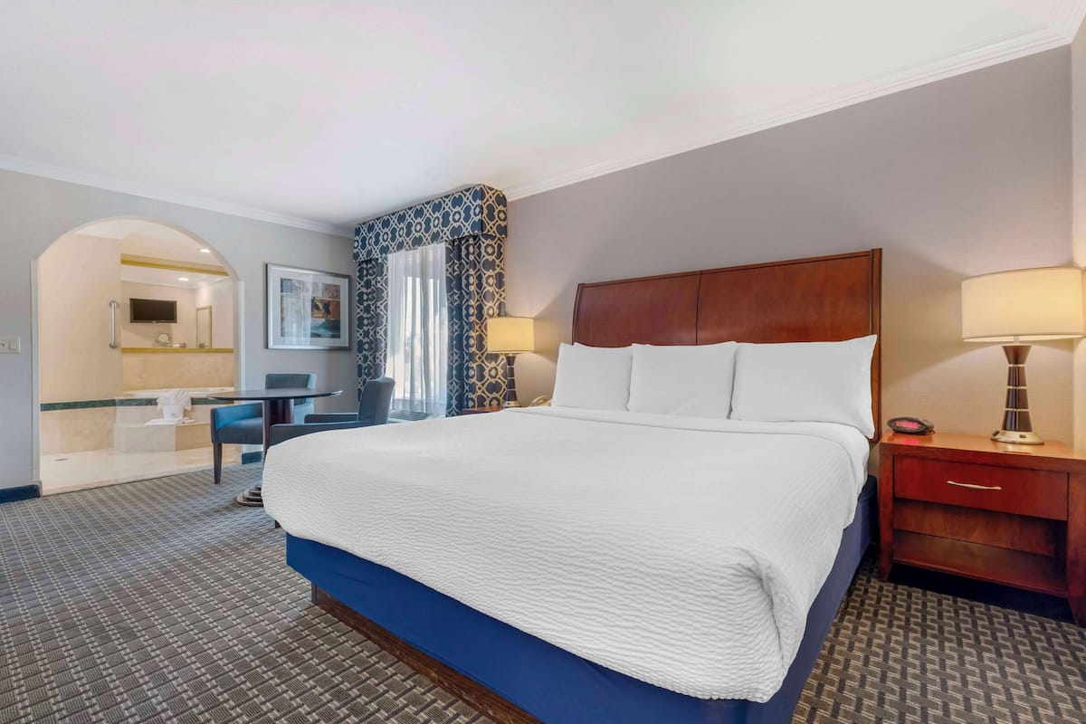 A hotel room with a large bed, a desk, and a chair. There is a mirror, a lamp, and a window with curtains.