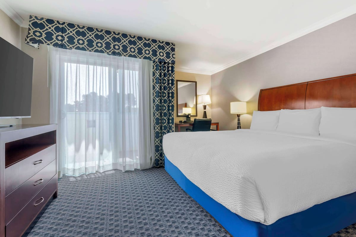A hotel room with a large bed, a desk with a lamp, a dresser, and a large window with sheer curtains.
