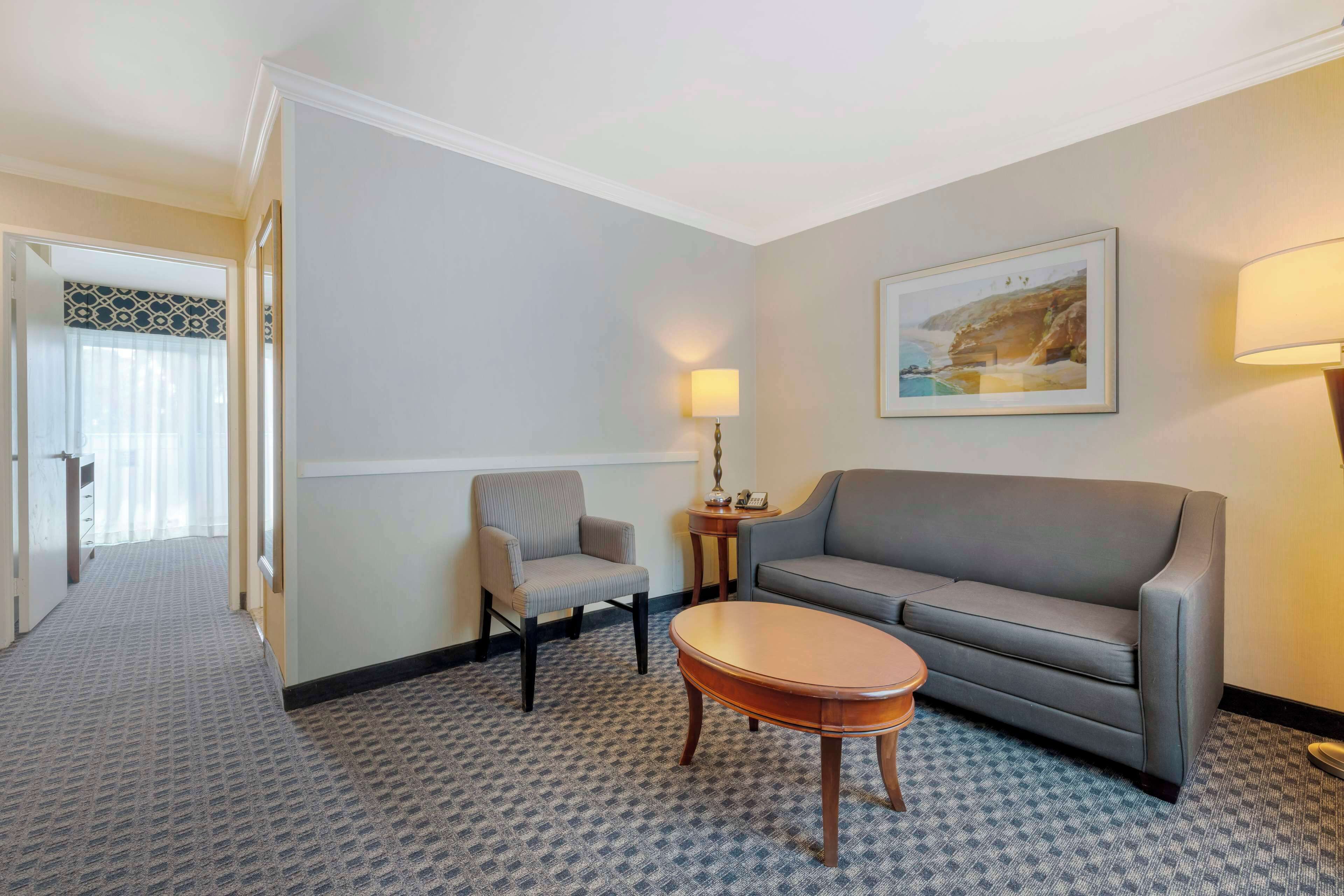 Een hotelkamer met een grijze bank, een fauteuil, een houten bijzettafeltje en een schilderij aan de muur.