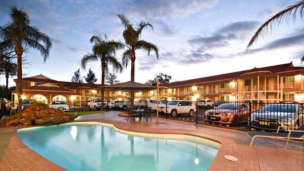A hotel with a swimming pool, palm trees, and a parking lot with cars. - The Aberdeen Motel (Dubbo)