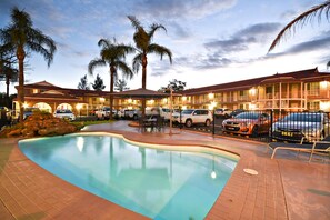 Un hôtel doté d’une piscine, de palmiers et d’un parking où sont garées des voitures.