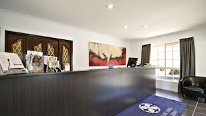 A hotel reception area with a dark wood reception desk, a large abstract painting, a blue carpet with a soccer ball logo, and a view of the outside through large windows. - The Aberdeen Motel (Dubbo)