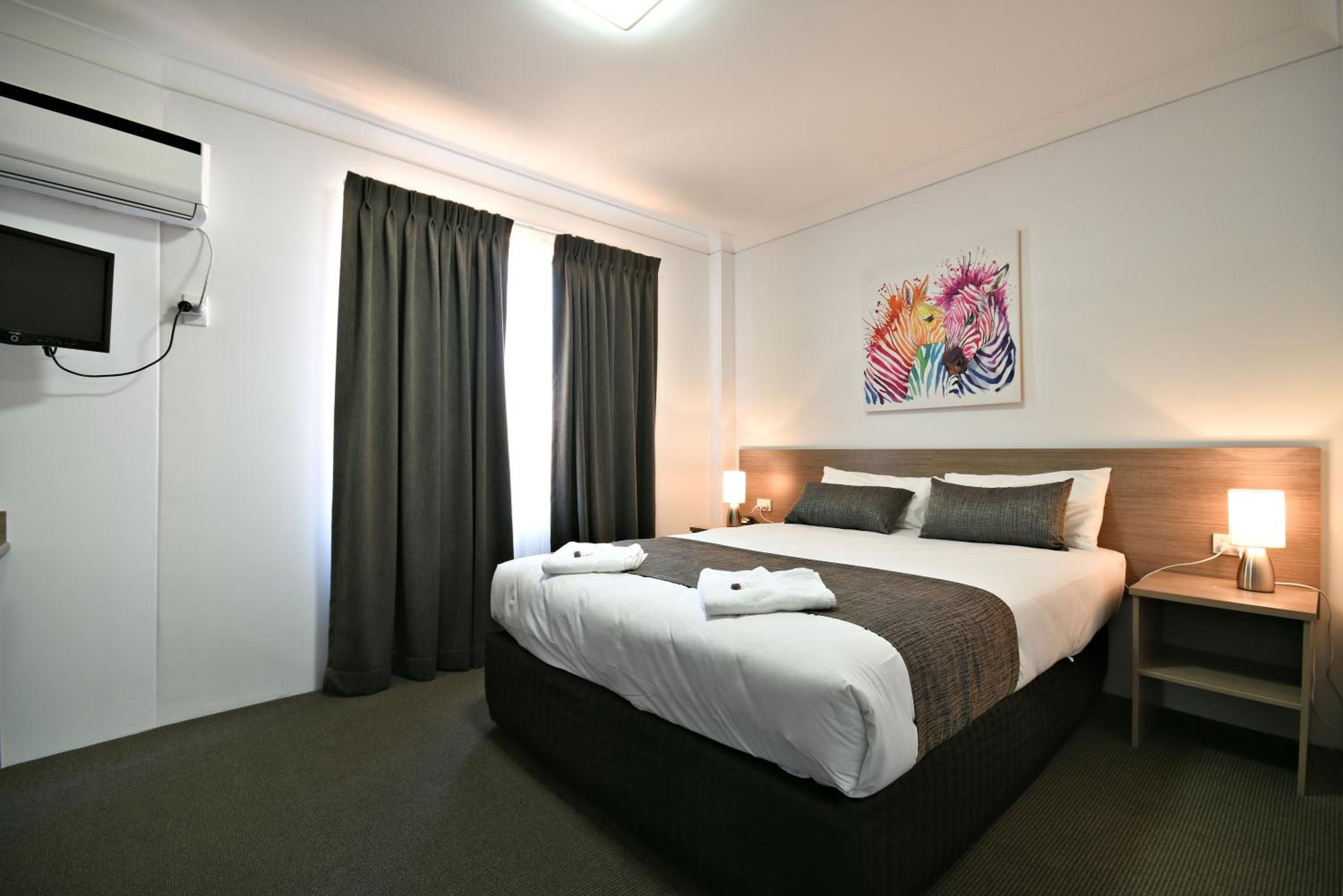 A hotel room with a bed, curtains, a flat-screen TV, a painting, and bedside lamps.