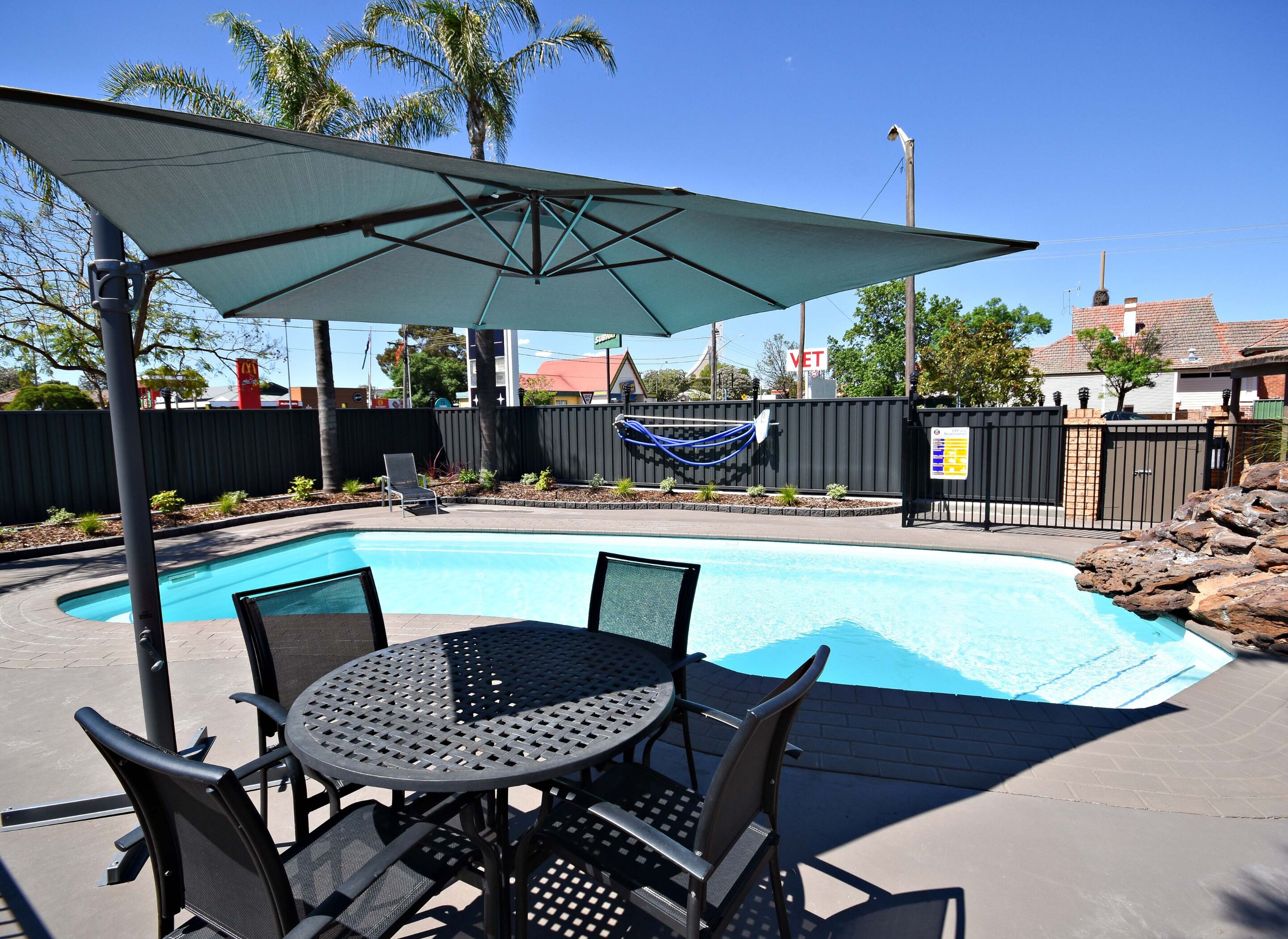 A pool area with a table and chairs, a covered umbrella, and a fenced yard with palm trees.