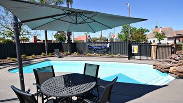 A pool area with a table and chairs, a covered umbrella, and a fenced yard with palm trees.