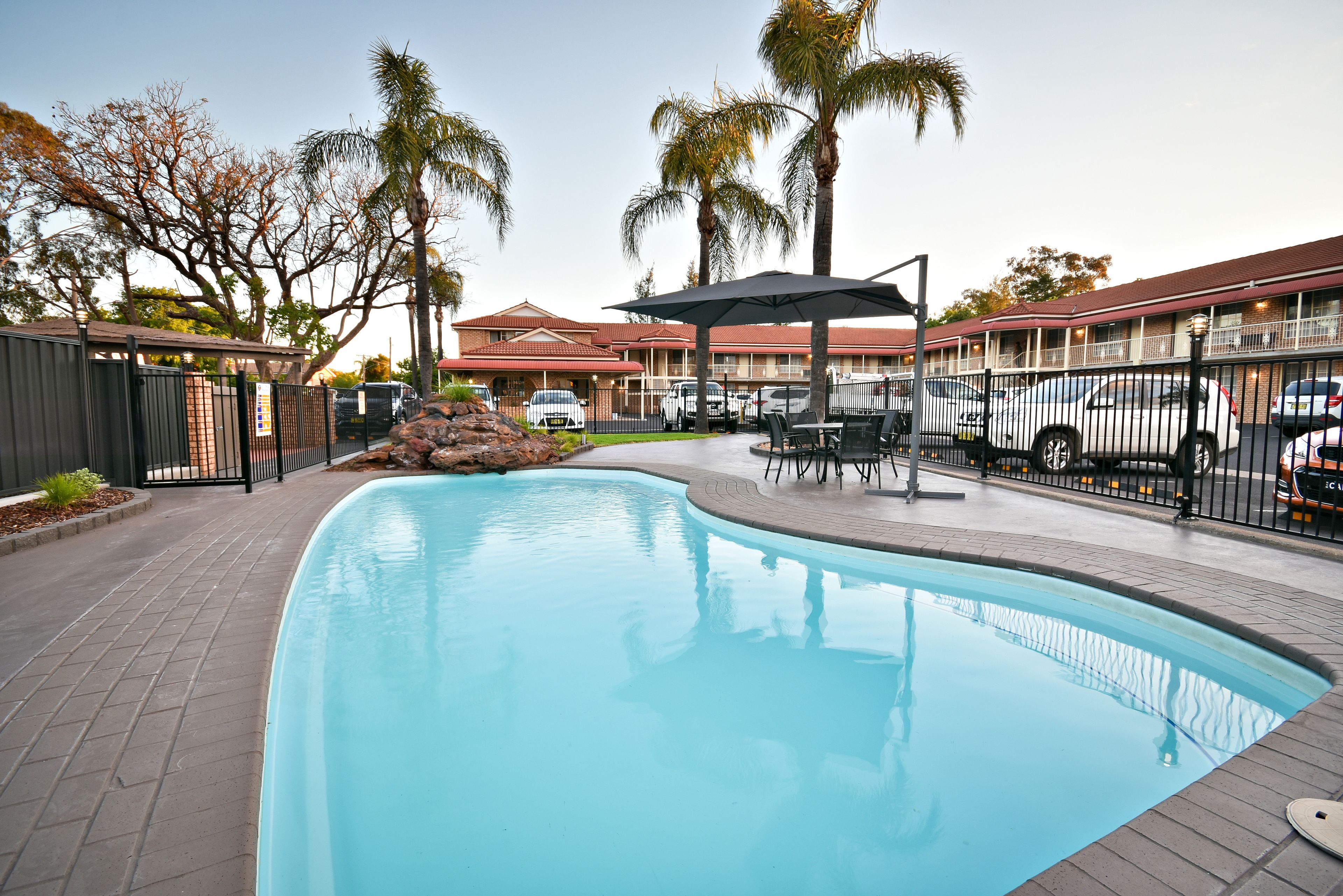 A swimming pool with a curved edge, surrounded by a paved walkway. There are palm trees and a building with a red roof in the background.