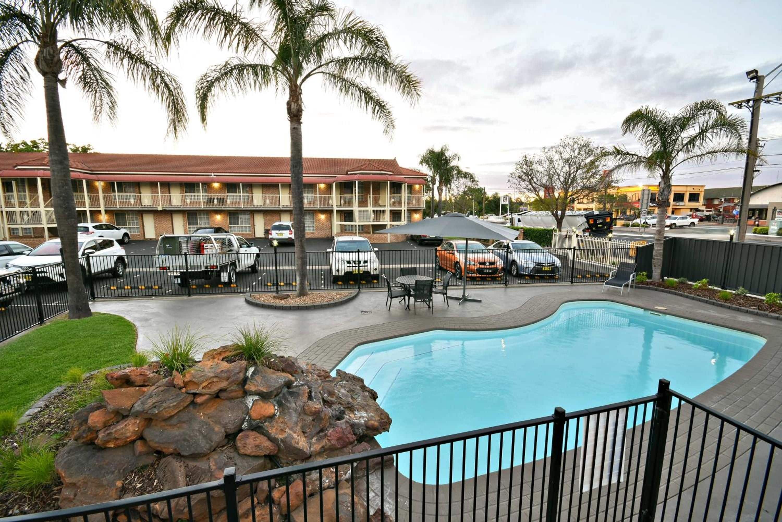 A swimming pool with a surrounding fence, palm trees, and a building with multiple balconies in the background.