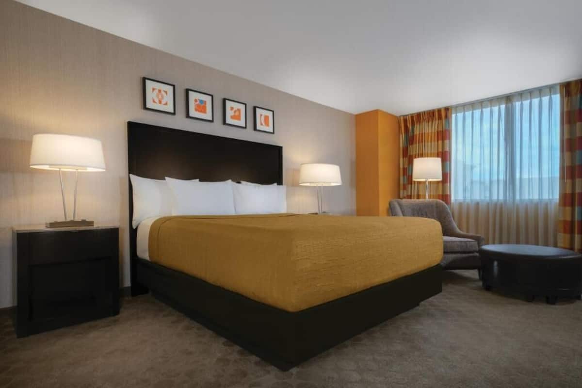 Ein Hotelzimmer mit einem großen Bett, zwei Nachttischen mit Lampen, einem Sessel, einem kleinen Hocker und einem Fenster mit Vorhängen.