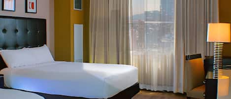 A hotel room with a large bed, a desk, and a view of the city through sheer curtains.