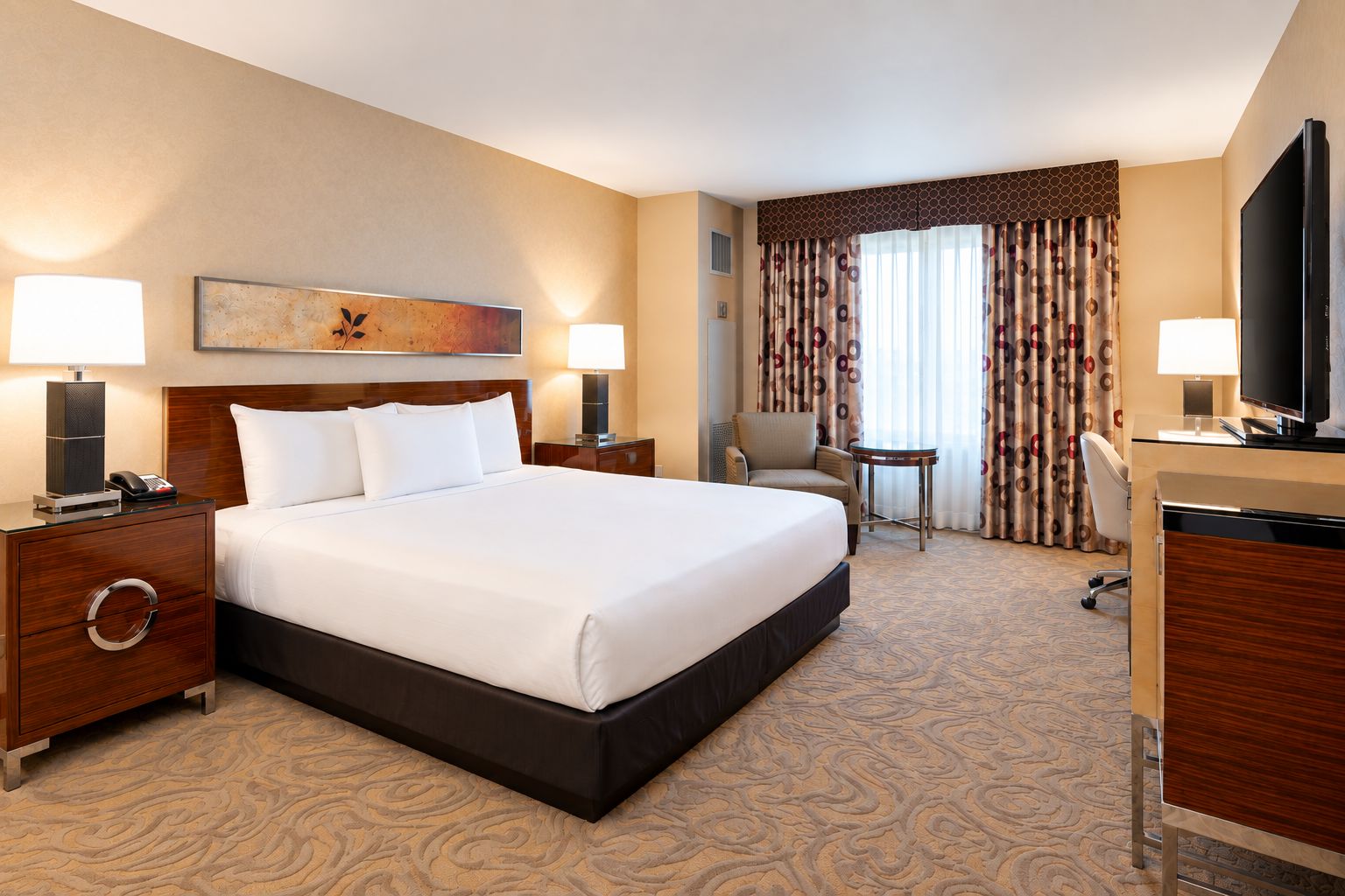 Une chambre d’hôtel avec un grand lit, deux tables de chevet avec des lampes, un bureau avec une télévision, une chaise et une fenêtre avec des rideaux.