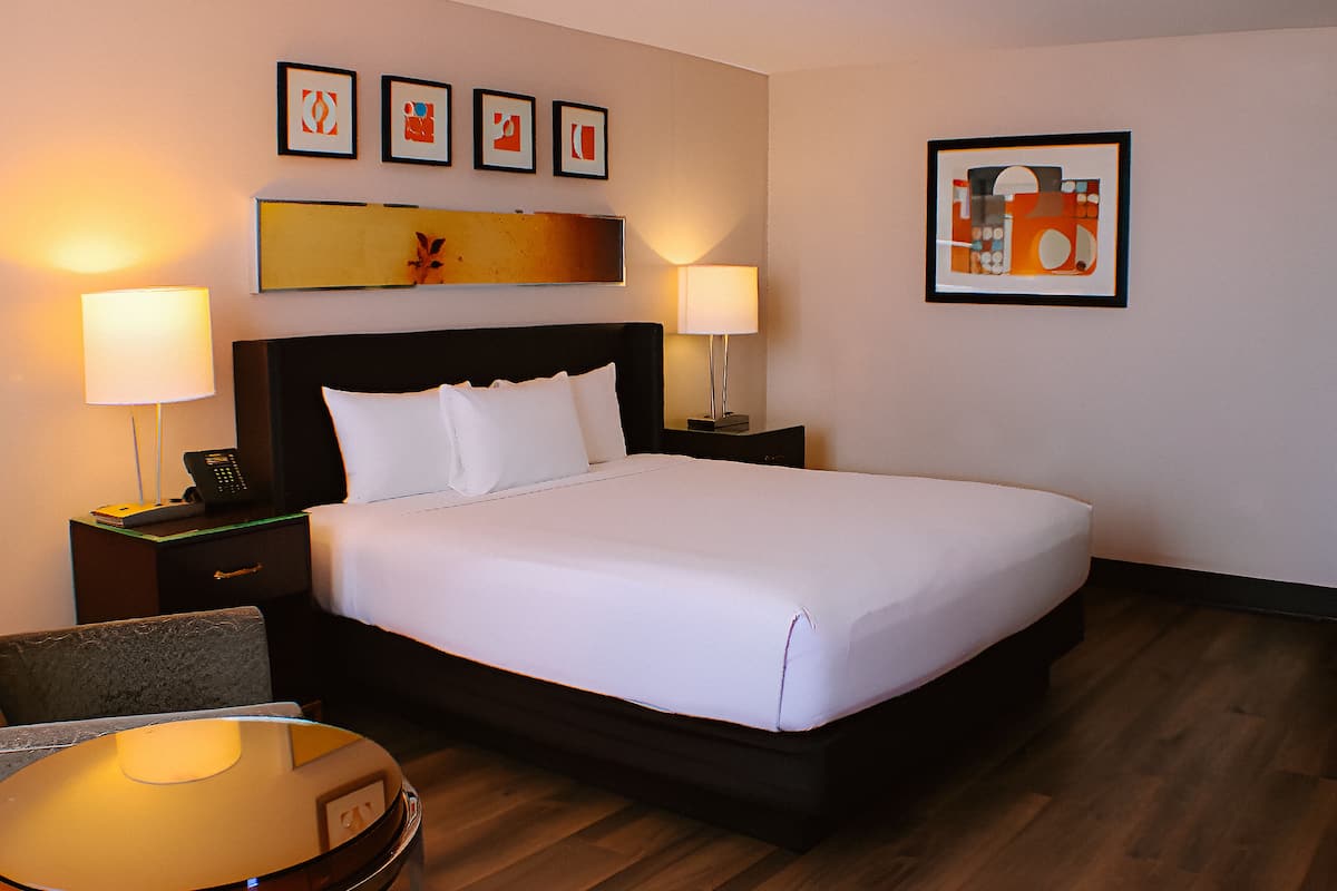 Ein Hotelzimmer mit einem großen Bett, zwei Nachttischen mit Lampen, einem Sessel und Kunstwerken an den Wänden.