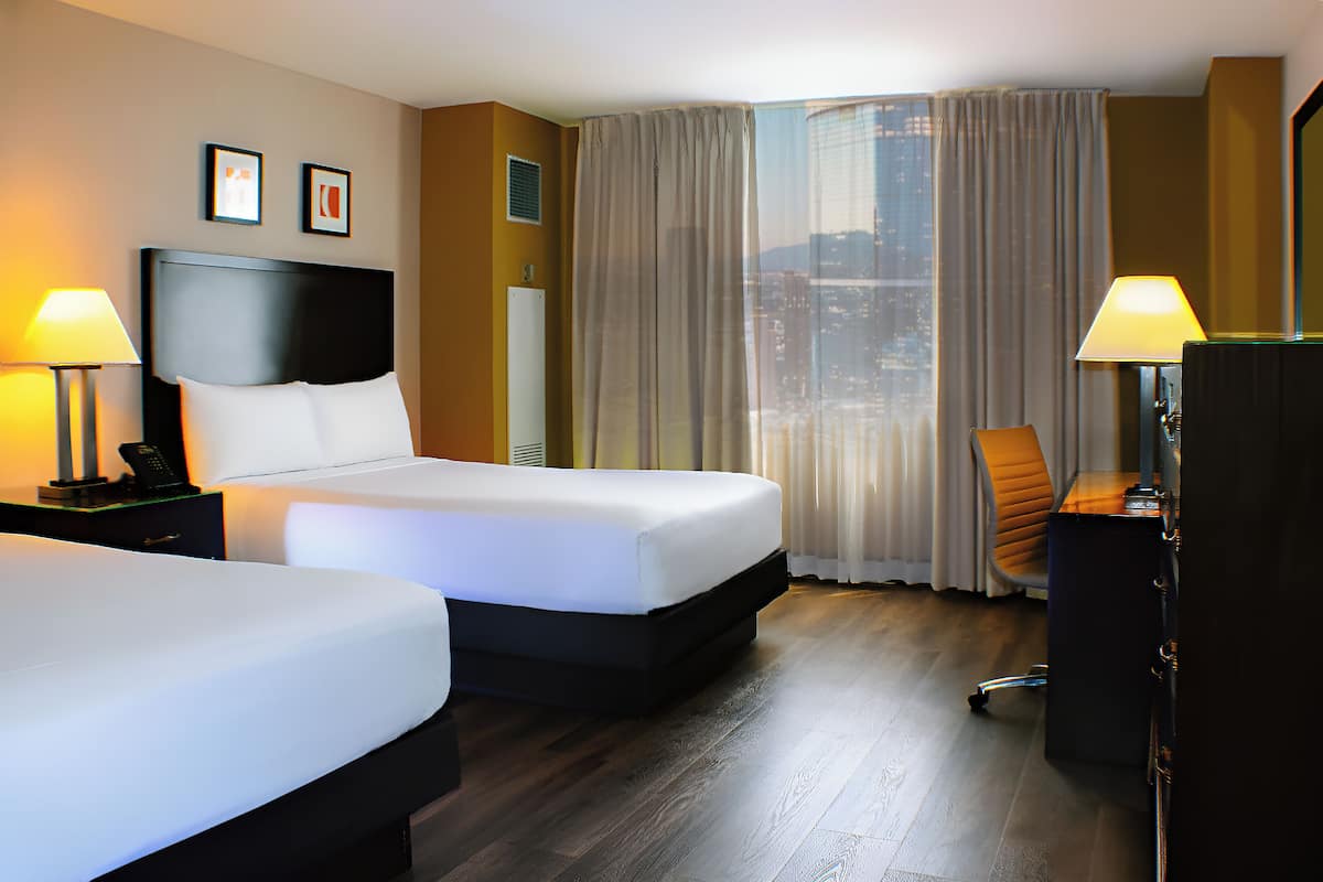 Ein Hotelzimmer mit zwei Betten, einem Schreibtisch und einem Stuhl. Auf den Nachttischen stehen je eine Lampe.