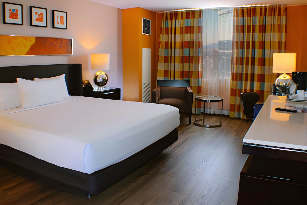 Ein Hotelzimmer mit einem großen Bett, einem Schreibtisch, zwei Stühlen, einem kleinen Tisch und Blick auf die Stadt durch Vorhänge.