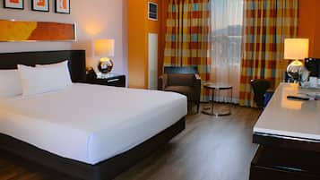 A hotel room with a large bed, a desk, two chairs, a small table, and a view of the city through curtains.