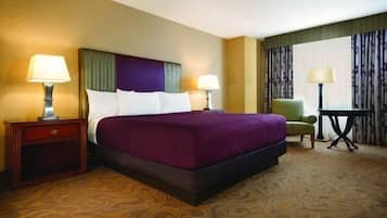 Quarto de hotel com uma cama grande, duas mesas de cabeceira com luminárias, uma poltrona verde e uma pequena mesa com luminária.
