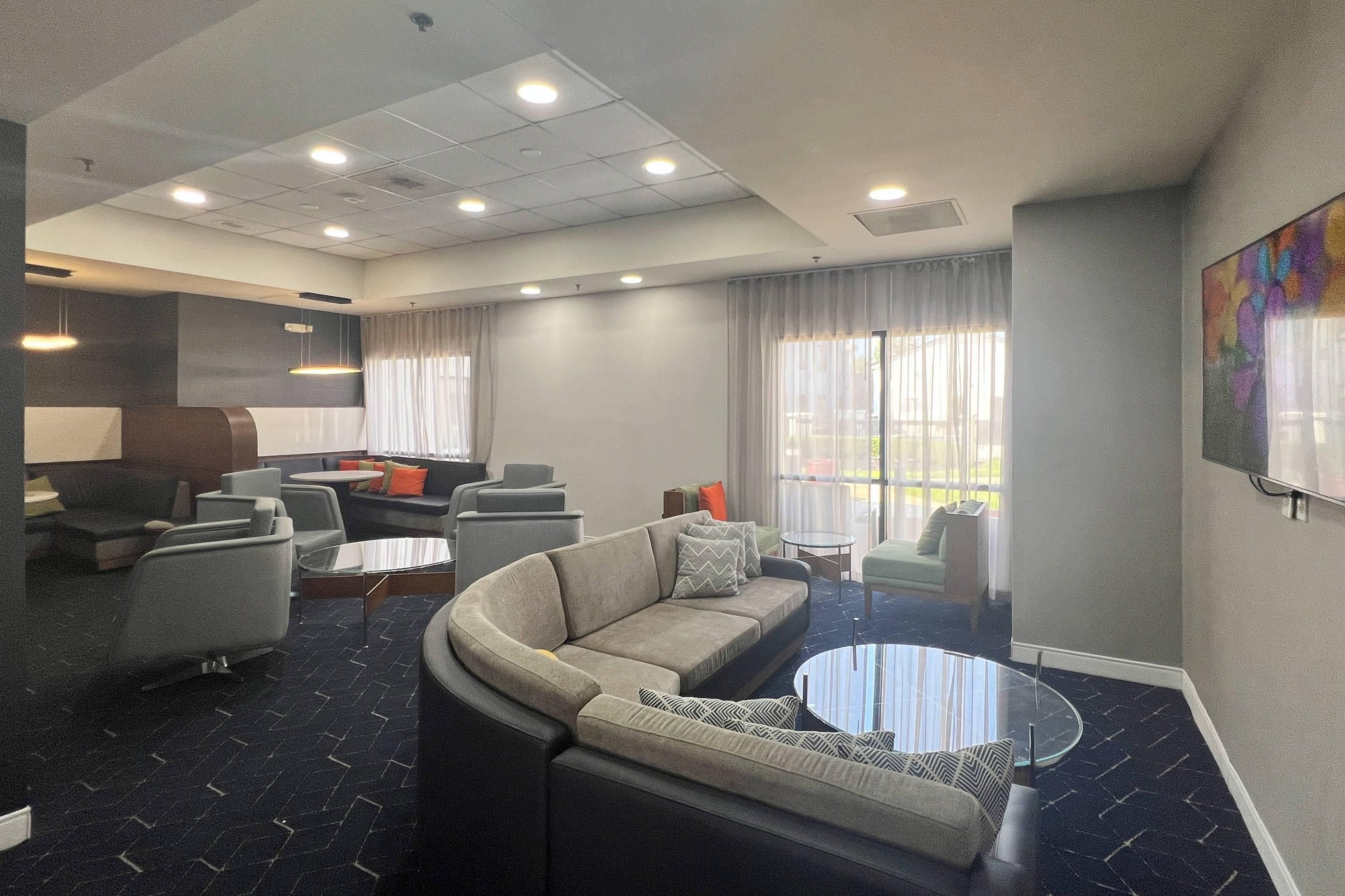 A modern hotel lobby with a curved sofa, armchairs, a coffee table, and a large window with curtains.