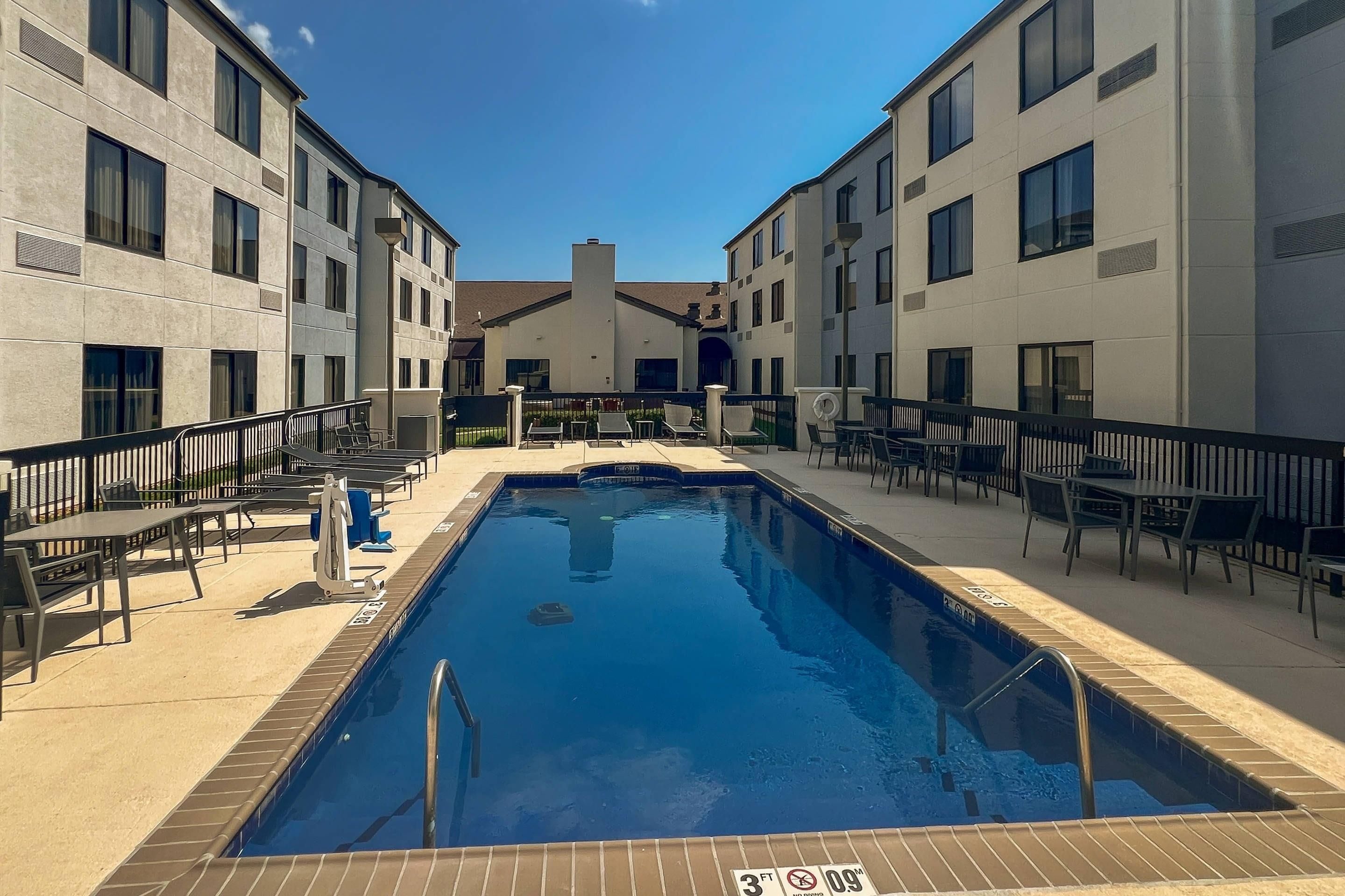 A rectangular swimming pool with a diving board and lounge chairs around it, surrounded by a building with multiple windows and balconies.
