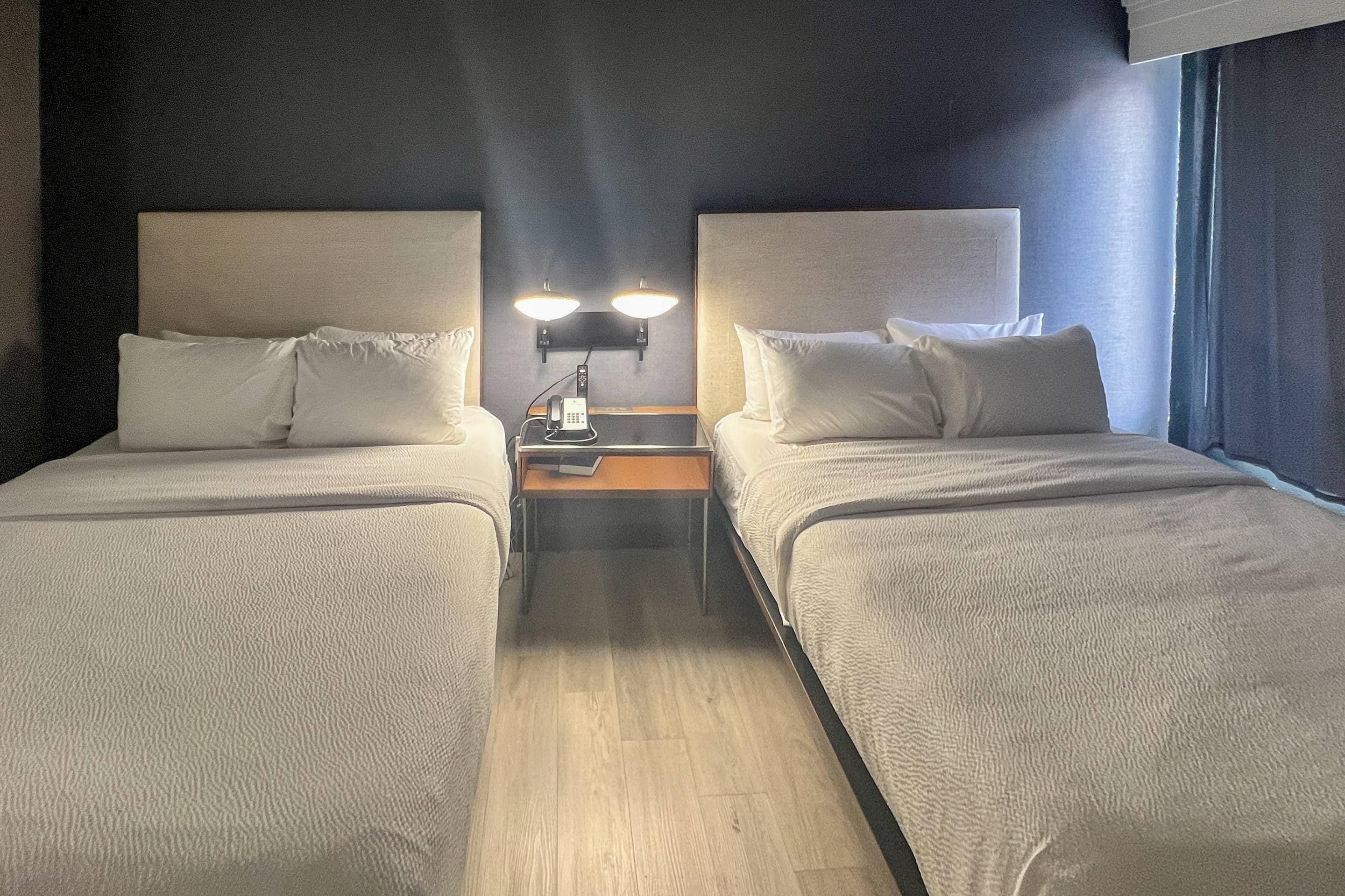 A hotel room with two beds, a nightstand with a phone, and a wall-mounted light fixture.
