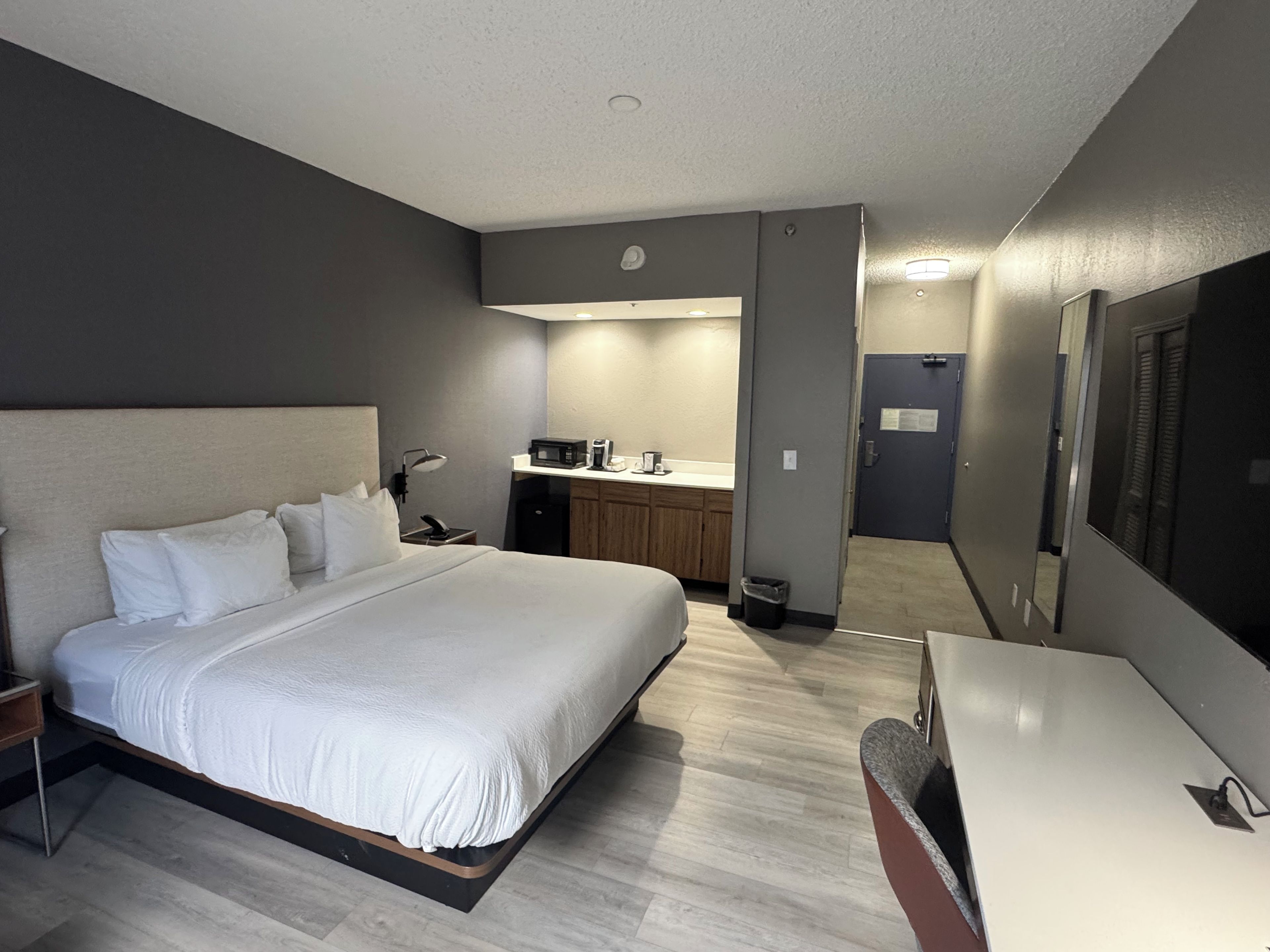 Een hotelkamer met een bed, nachtkastje, lamp, en een kitchenette met een magnetron en koffiezetapparaat.