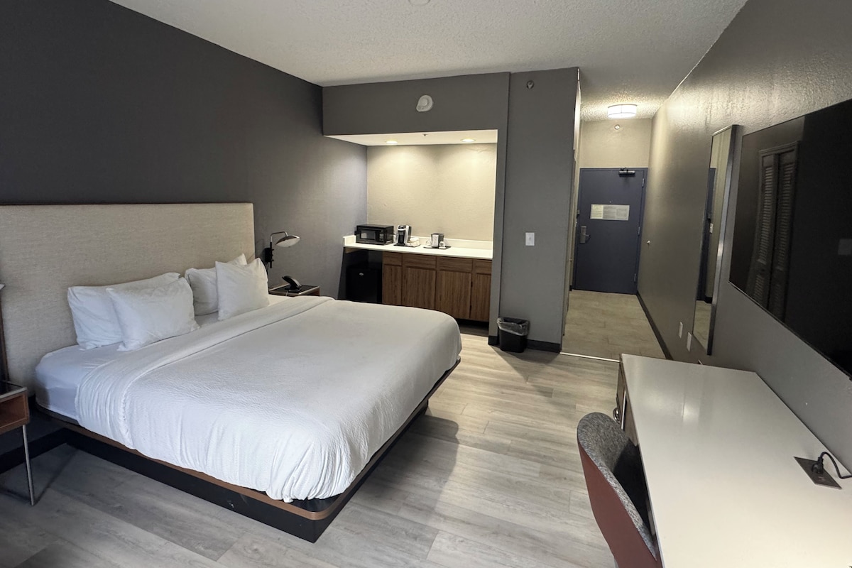 Een hotelkamer met een bed, nachtkastje, lamp, en een kitchenette met een magnetron en koffiezetapparaat.