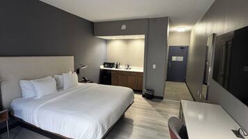 Een hotelkamer met een bed, nachtkastje, lamp, en een kitchenette met een magnetron en koffiezetapparaat.